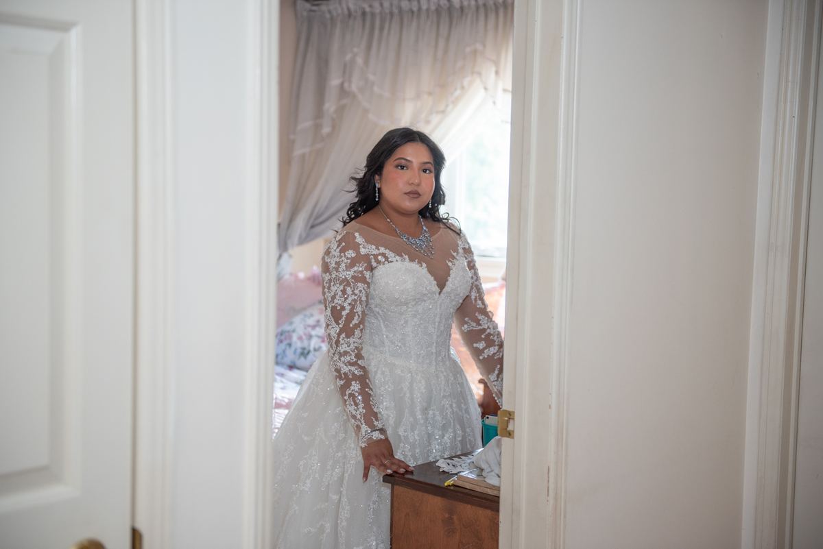 A bride in a white lace gown stands pensively in a bedroom, illuminated by soft light from an open window.