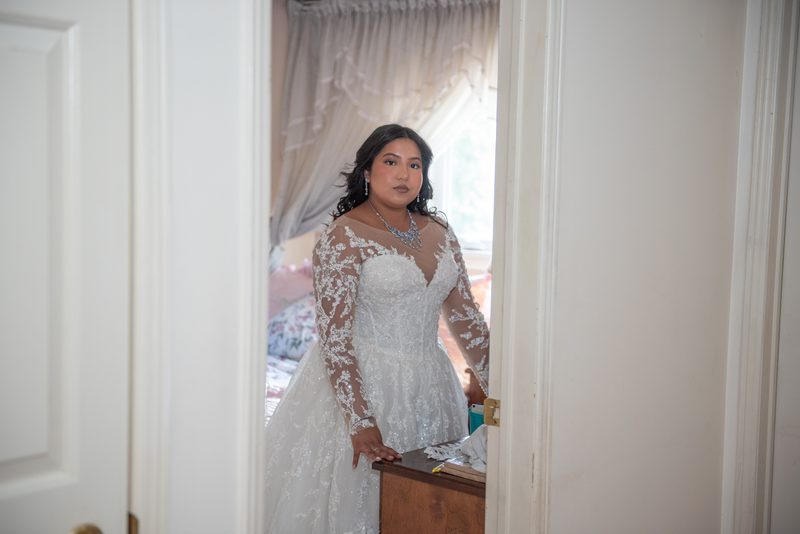 A bride in a white lace gown stands pensively in a bedroom, illuminated by soft light from an open window.