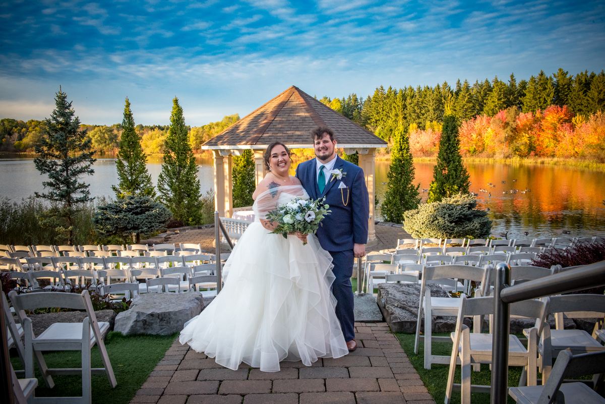 A smiling newlywed couple stands on a paved aisle by a lake, framed by white chairs and a wooden gazebo.