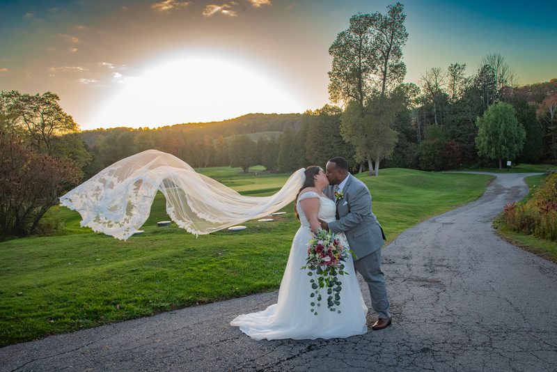 A newlywed couple kisses beside a golf course, the bride's veil billowing in the wind during a golden sunset.