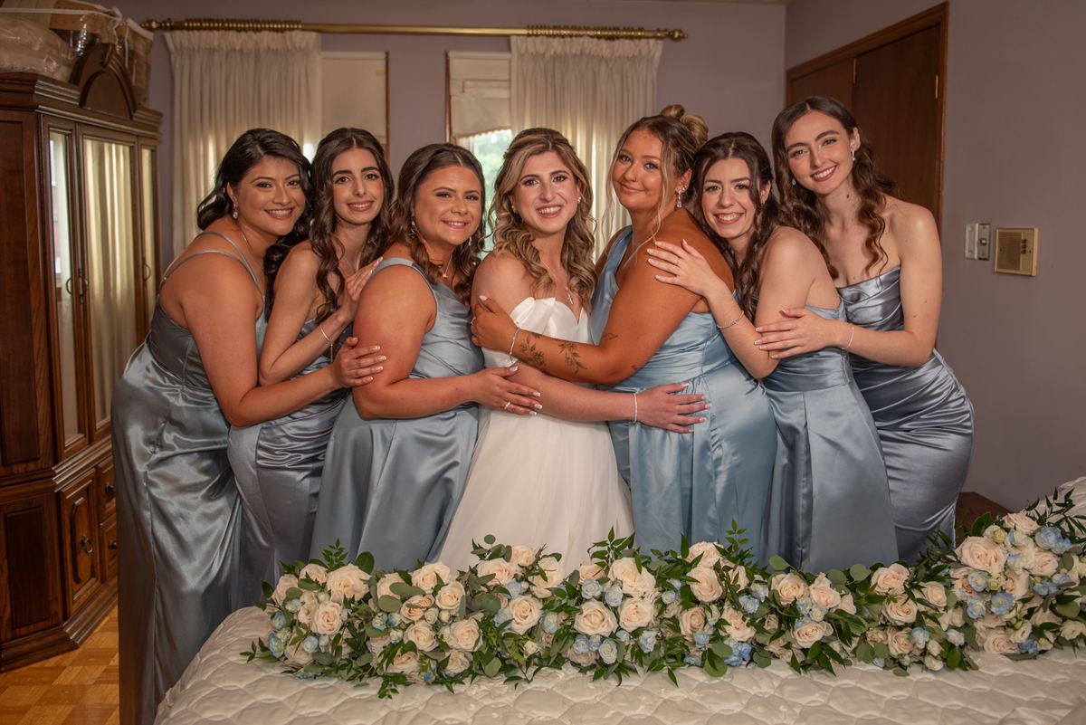 Bride in white gown embraced by smiling bridesmaids in blue dresses near bed with floral arrangement.