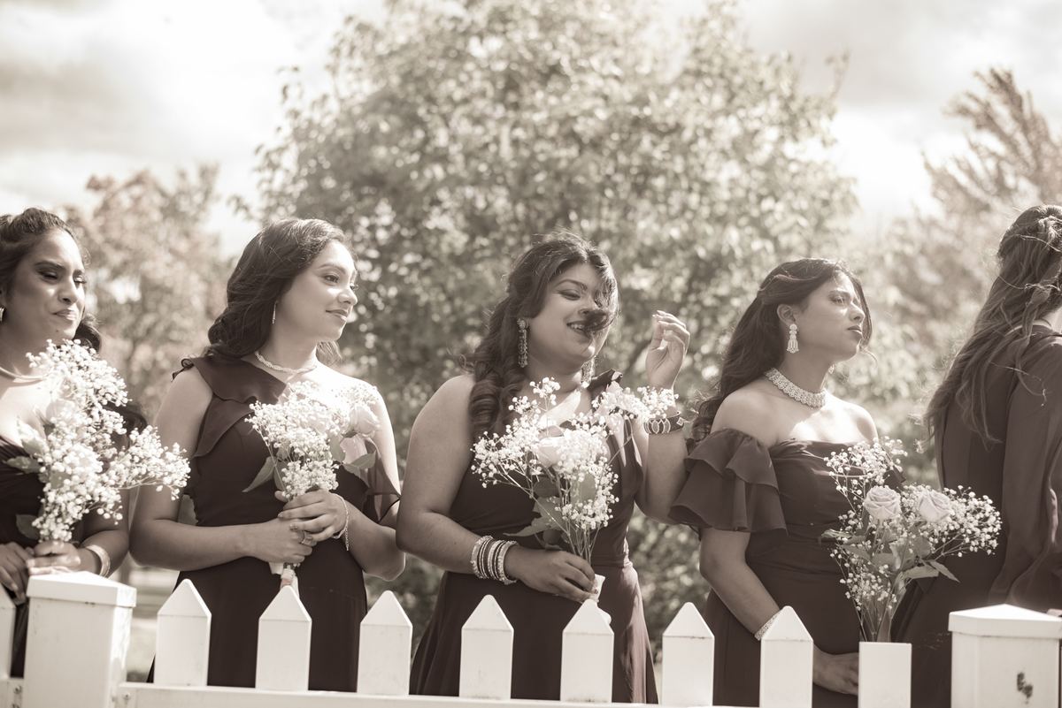 Bridesmaids in dark gowns and jewelry hold bouquets behind a white fence, watching with serene expressions.