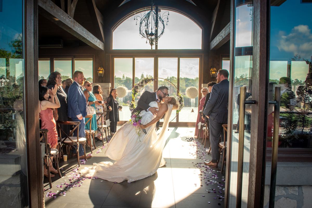 A bride and groom kiss at their wedding, surrounded by guests and golden sunlight pouring through arched windows.