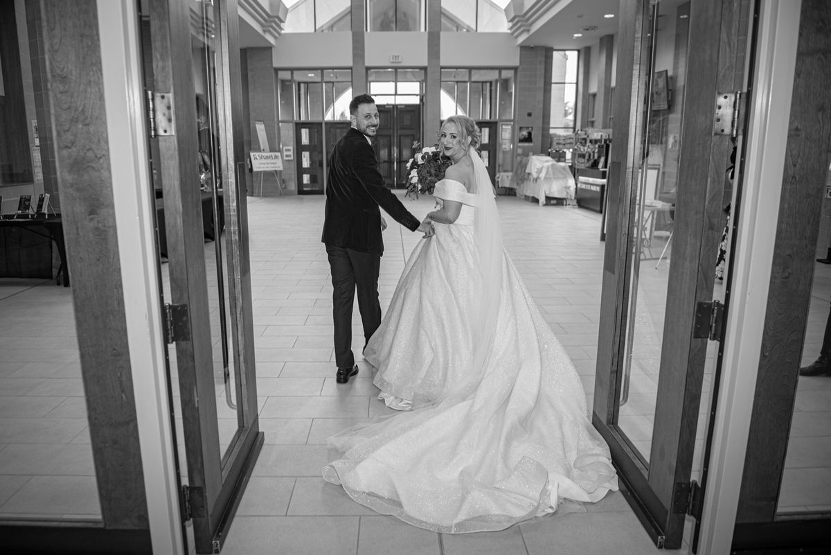Bride in sparkling gown and groom enter modern hall through glass doors, she turns to smile at camera.