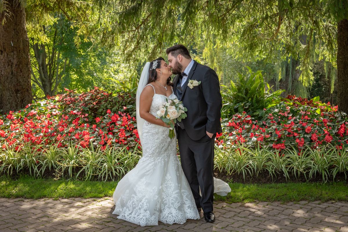 A bride and groom kiss in a sunlit garden surrounded by green trees and red flowers, illuminated by dappled sunlight.