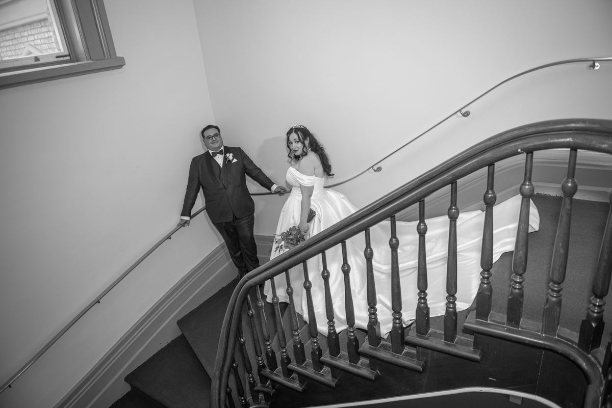 A smiling bride and groom descend a wooden staircase, their attire highlighted by soft light from an unseen window.