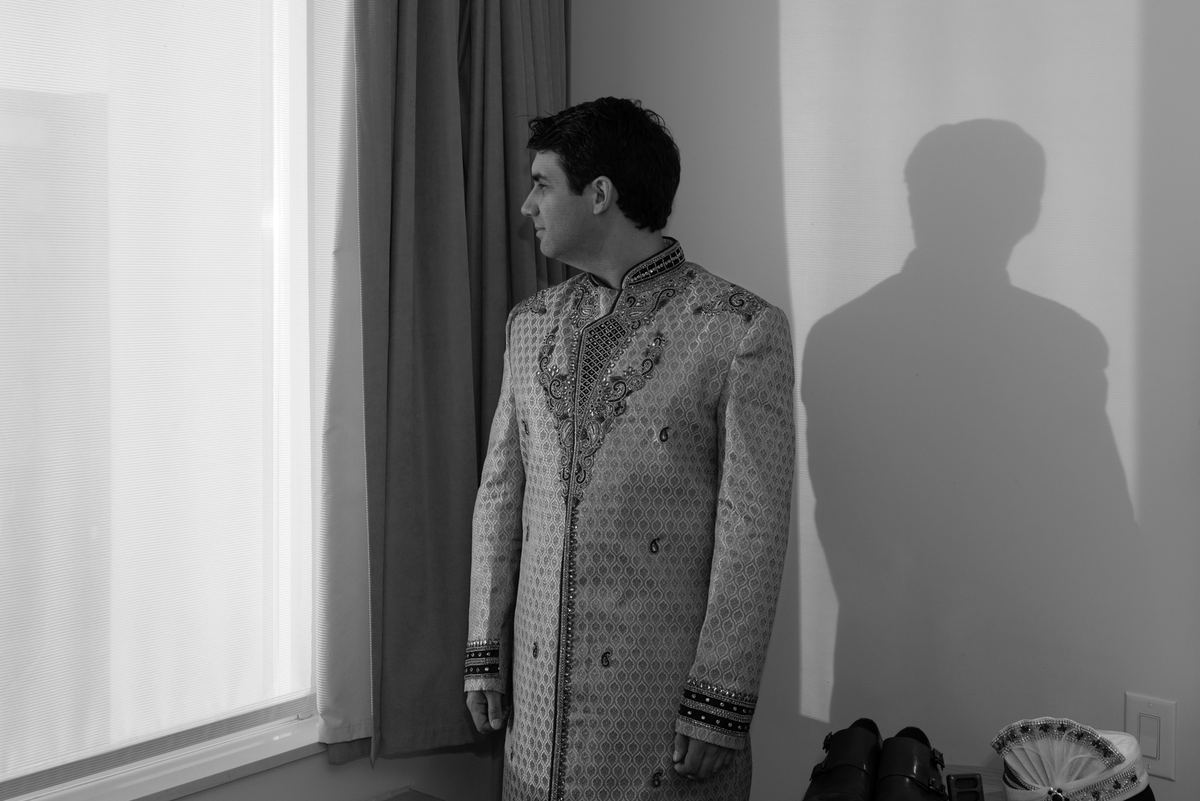 A man in an ornate sherwani stands by a window, illuminated by soft light casting a shadow on the wall.