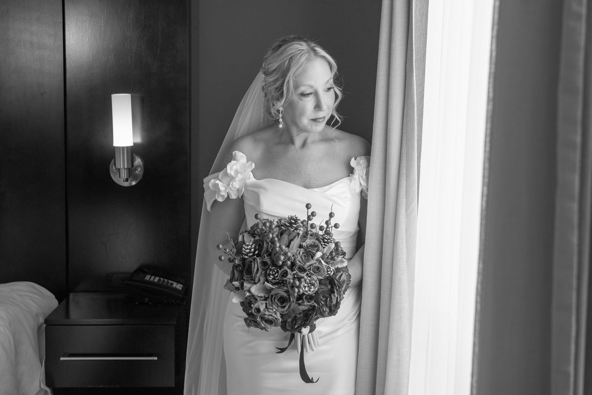 A bride in a wedding gown and veil holds a bouquet, gazing thoughtfully out a window in a softly lit room.