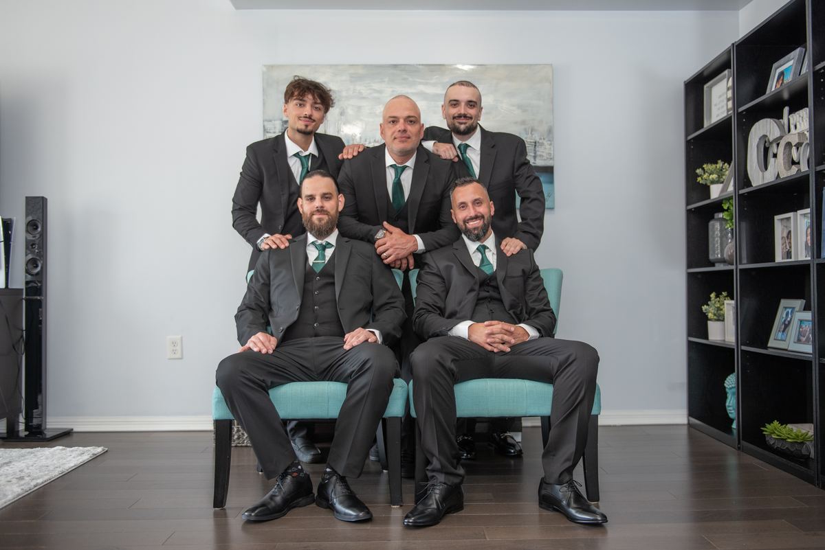 Five formally dressed men pose together in a modern living room, smiling and looking toward the camera.