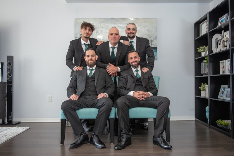 Five formally dressed men pose together in a modern living room, smiling and looking toward the camera.