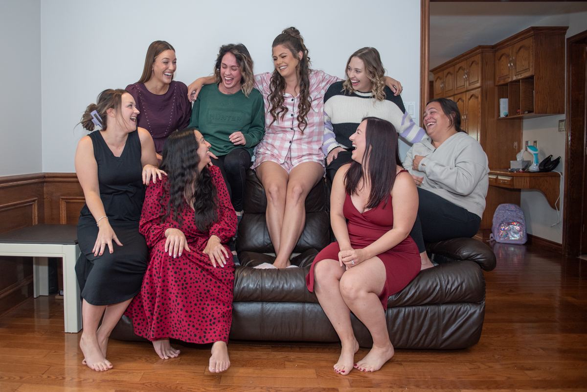 Seven women laughing together on a brown leather couch in a warm living space, their joyful expressions.