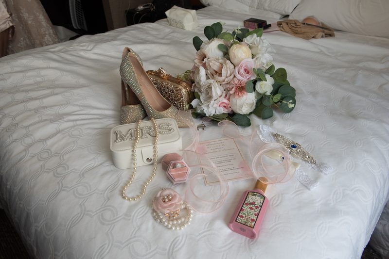 High heels, pearl necklace, bouquet, clutch, perfume bottle, and invitation arranged on white bedspread.
