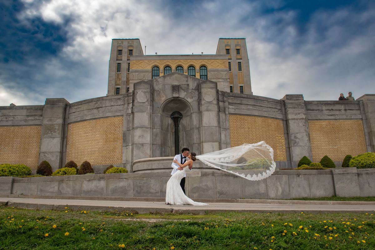 A bride and groom kiss in front of an Art Deco building, with grass and wildflowers in the foreground.