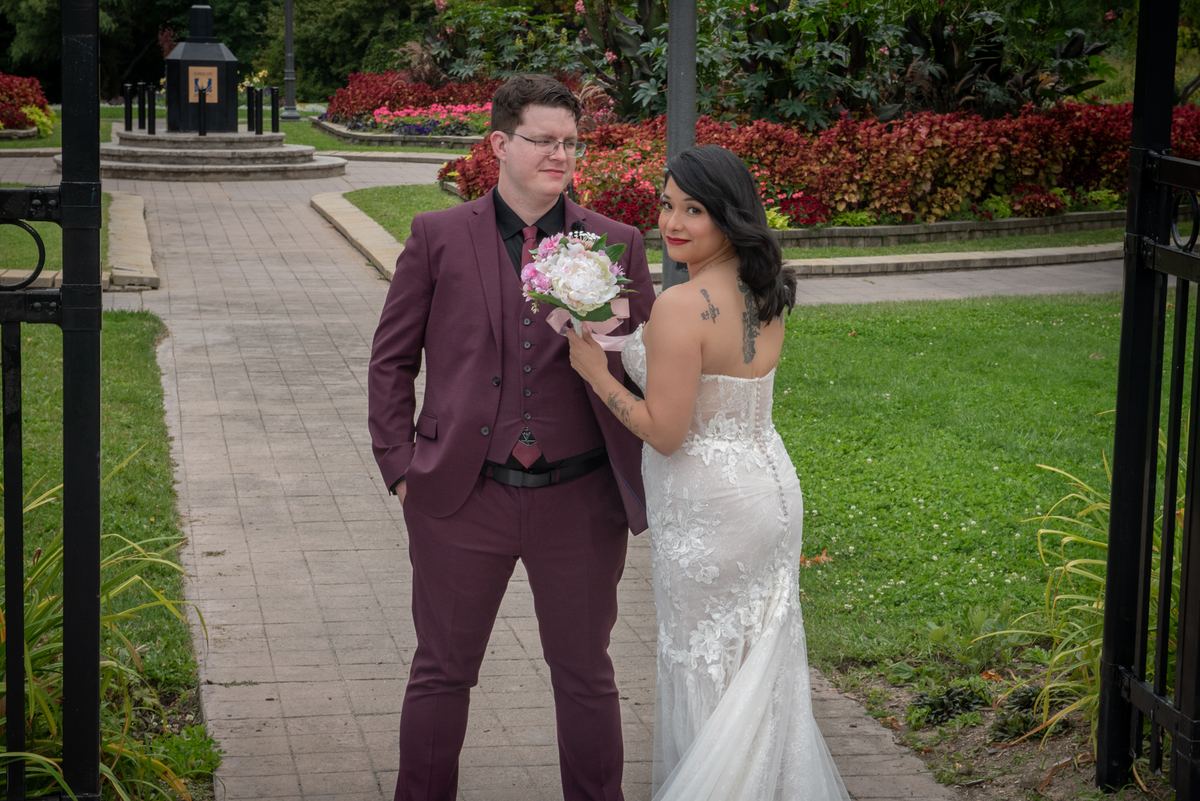 A bride in white and groom in burgundy stand on a garden path, smiling at each other amid lush greenery.