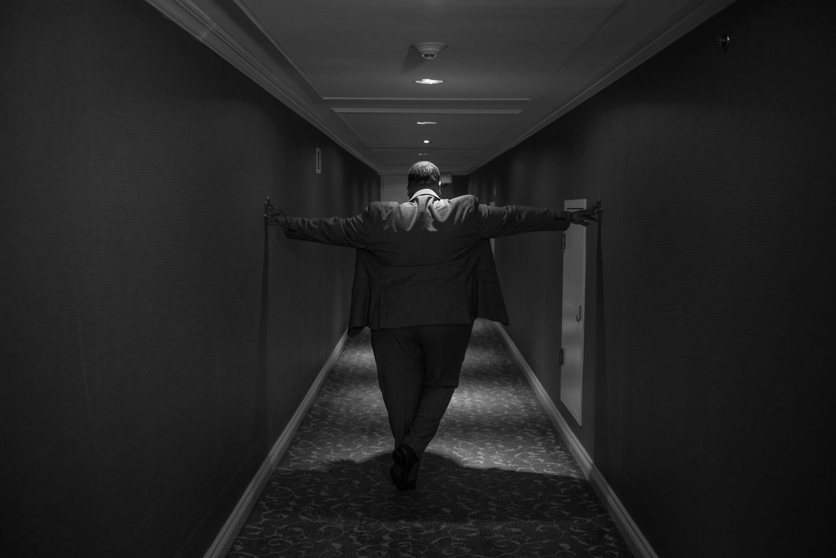 A man in a suit walks down a dimly lit corridor, arms outstretched, his long shadow emphasizing solitude.