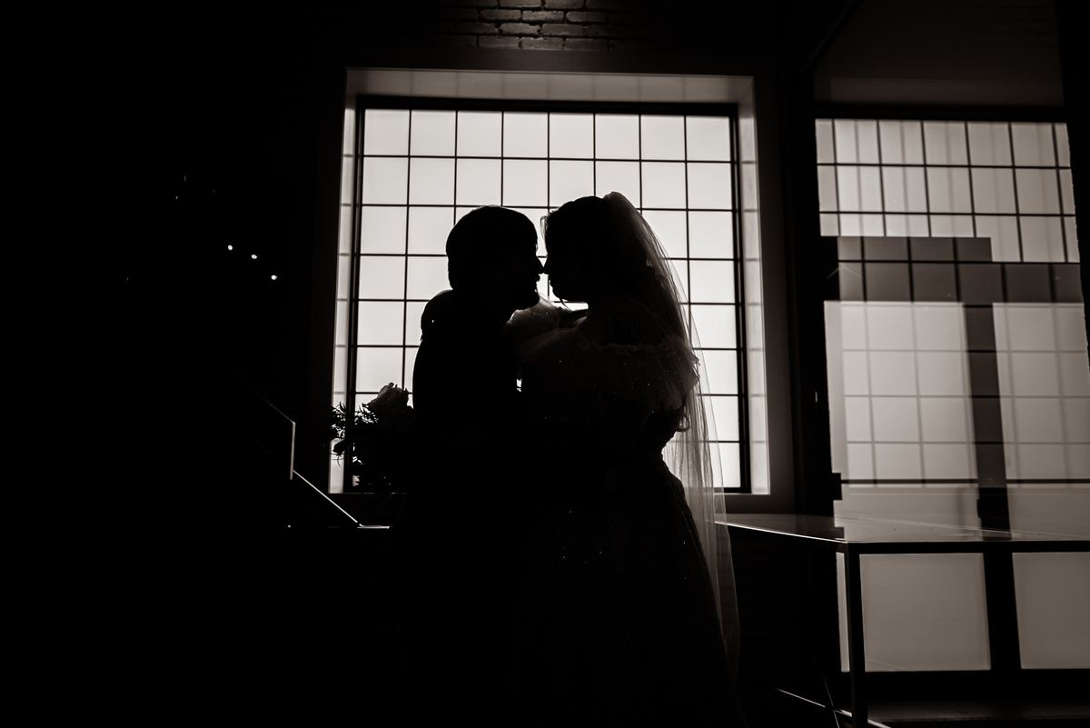 Bride and groom kiss in silhouette against a bright window grid, emphasizing their forms and the texture of her veil.