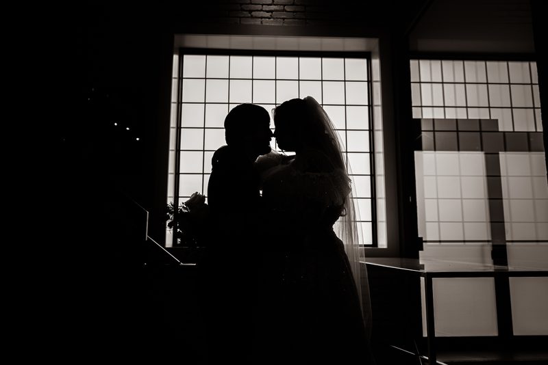 Bride and groom kiss in silhouette against a bright window grid, emphasizing their forms and the texture of her veil.