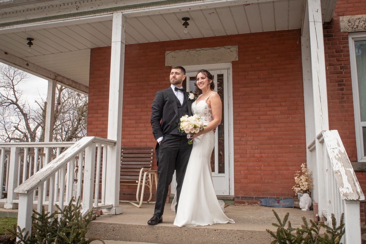 A smiling bride in white and groom in black tuxedo stand on a rustic porch with weathered railings, looking at the camera.