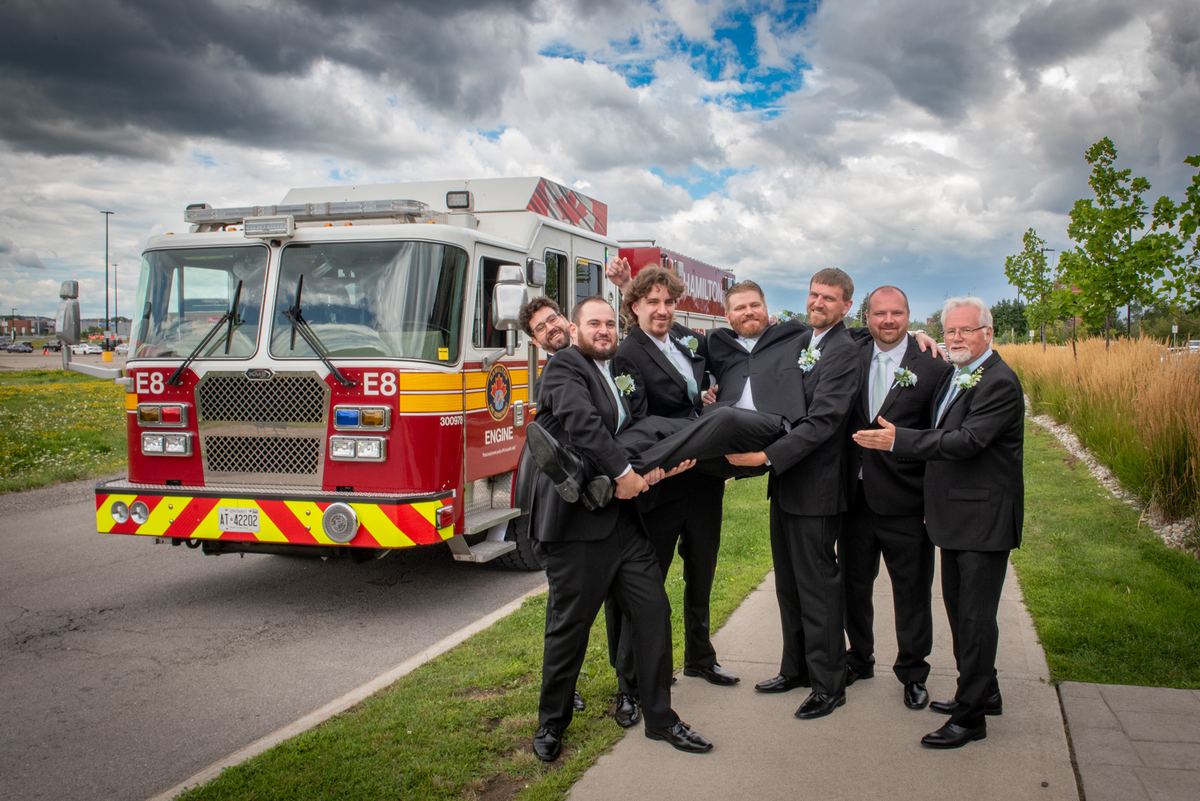 Groomsmen in black suits lift the groom beside a red fire engine under a partly cloudy sky with sunlight patches.