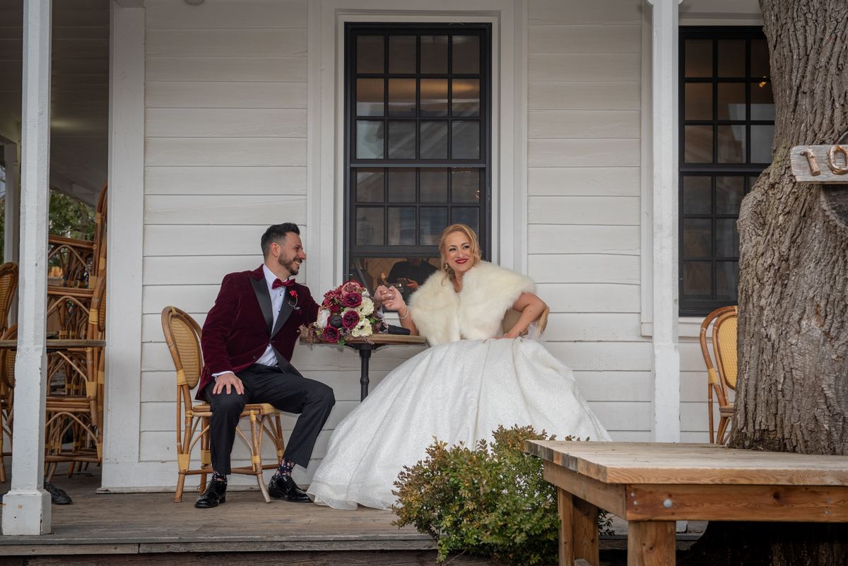 A newlywed couple in formal attire smiles and looks at each other on a porch, illuminated by natural light with floral decor.