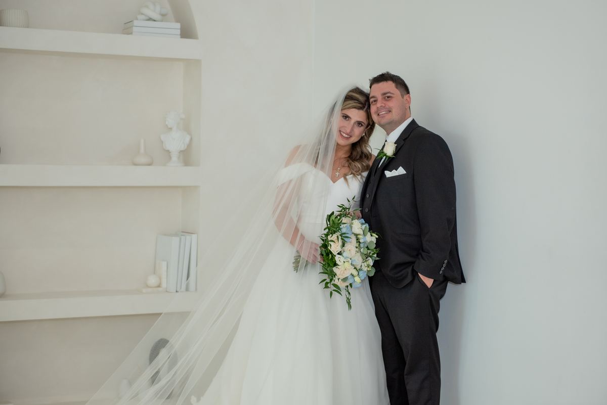 A bride and groom stand beside a white shelving unit, smiling warmly in a minimalist setting with pastel flowers.