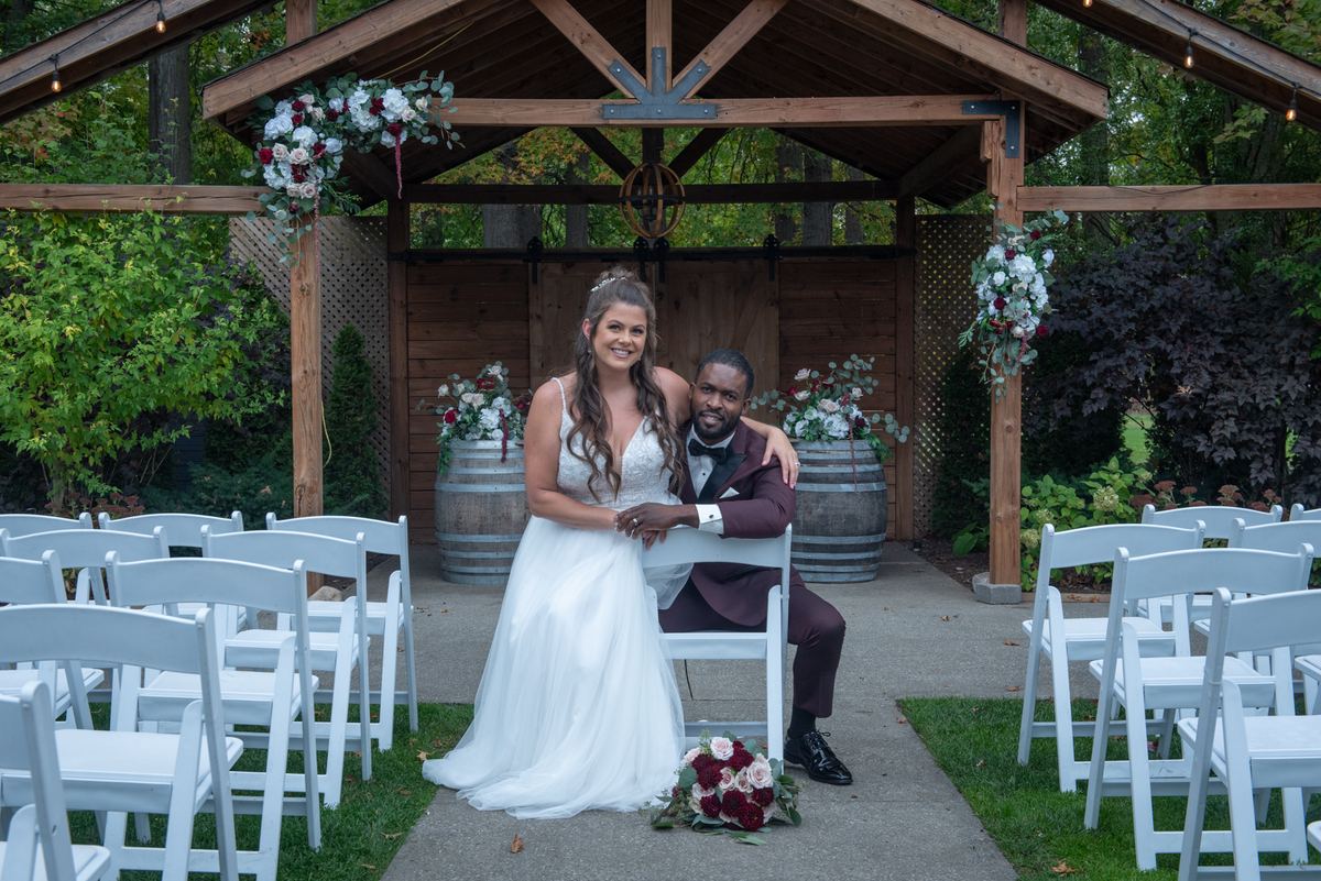A bride in a white gown and groom in a burgundy suit smile under a floral-decorated wooden arbor during an outdoor ceremony.