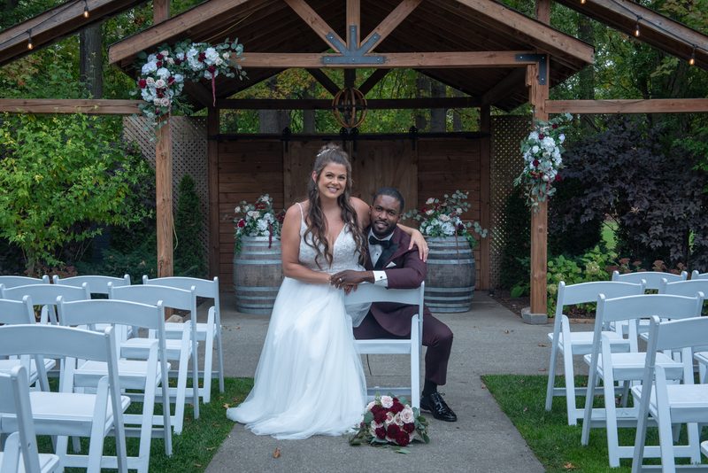 A bride in a white gown and groom in a burgundy suit smile under a floral-decorated wooden arbor during an outdoor ceremony.