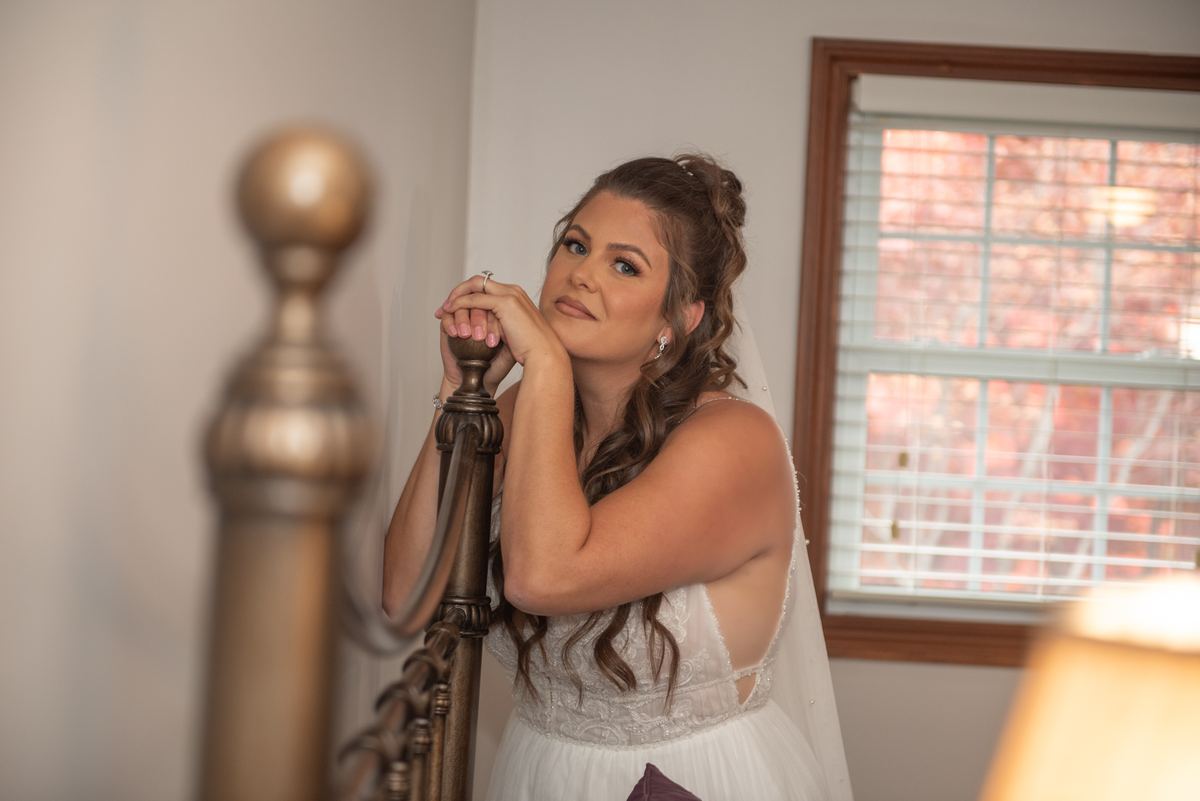 A bride in her wedding gown leans against a staircase railing, gazing upward with a serene expression.