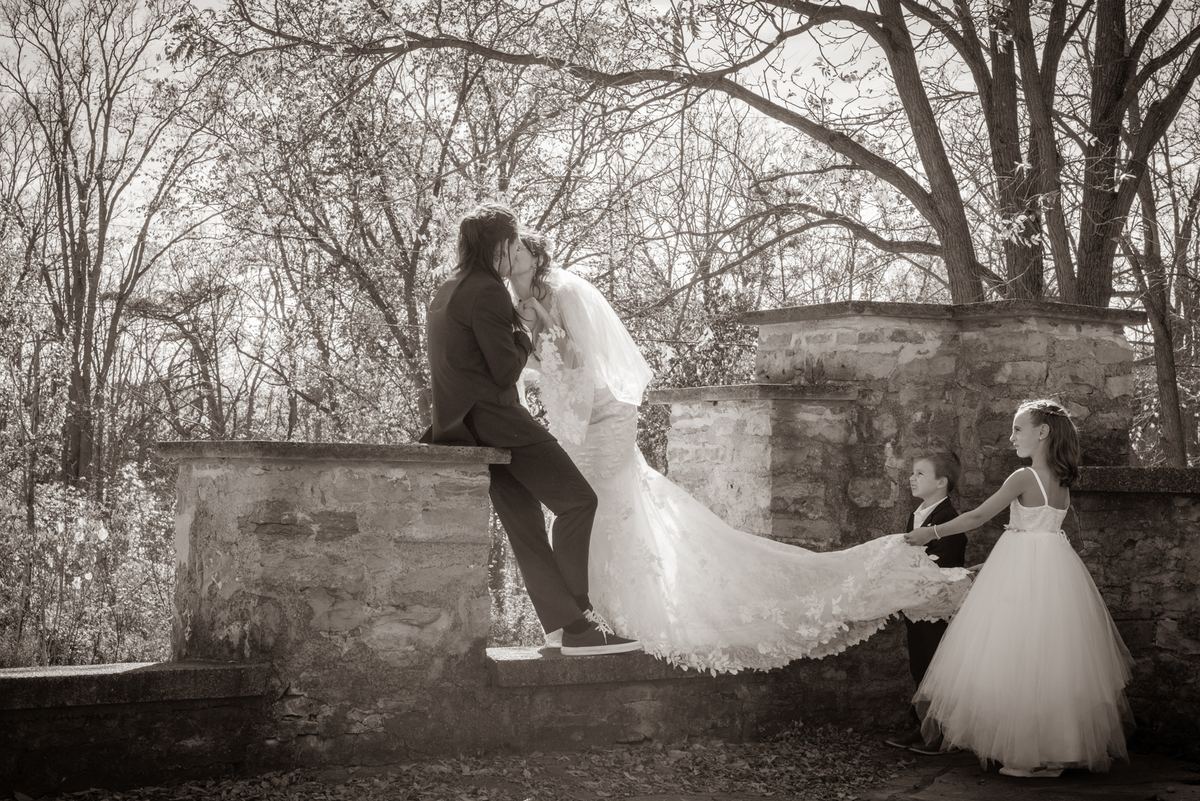 A bride and groom kiss while seated on a stone wall, with flower girls nearby in a softly lit, tree-lined setting.