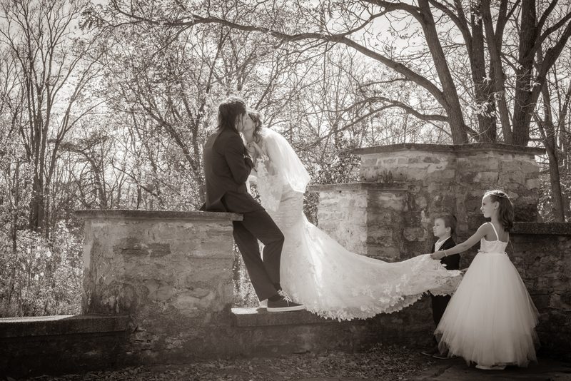 A bride and groom kiss while seated on a stone wall, with flower girls nearby in a softly lit, tree-lined setting.