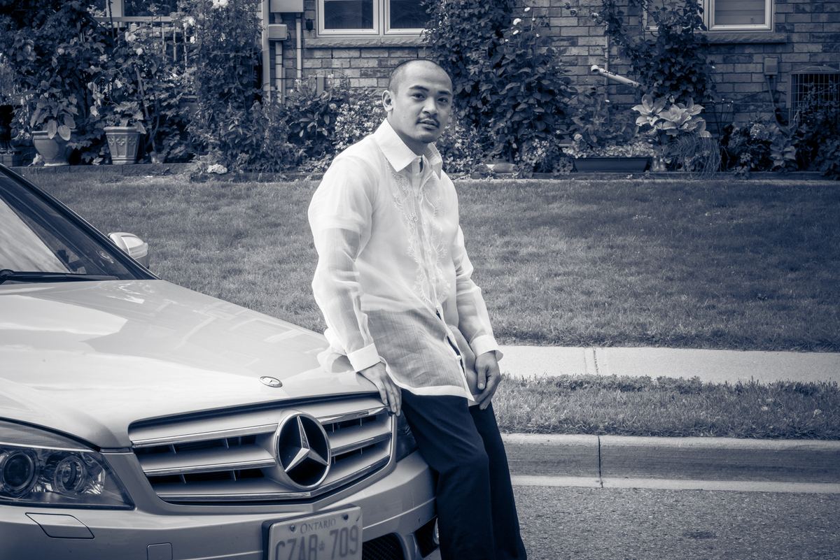 A man stands confidently against the hood of a mercedes-benz in a suburban neighborhood, wearing a crisp formal shirt.