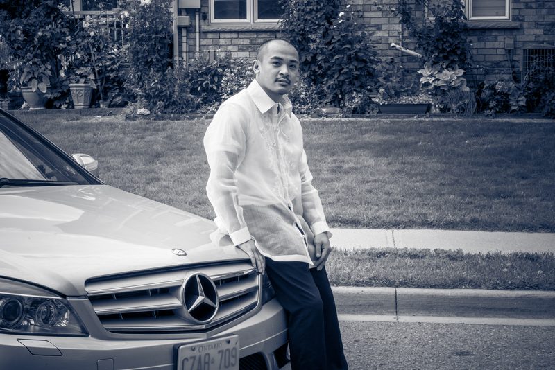 A man stands confidently against the hood of a mercedes-benz in a suburban neighborhood, wearing a crisp formal shirt.