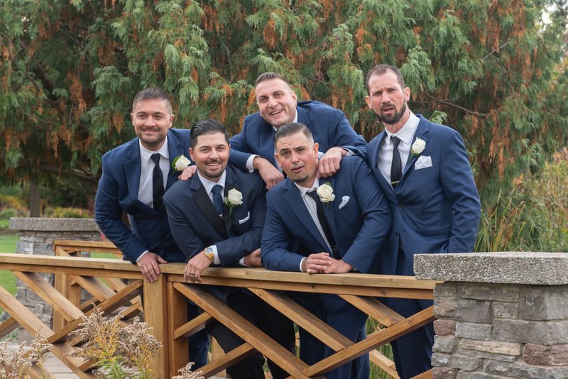 Five men in navy suits with white roses pose playfully on a wooden bridge surrounded by autumn trees.
