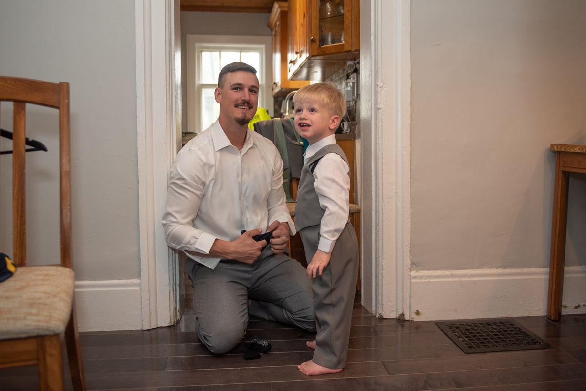 A groom kneels in a doorway adjusting his belt buckle while a young boy in matching attire stands beside him smiling.