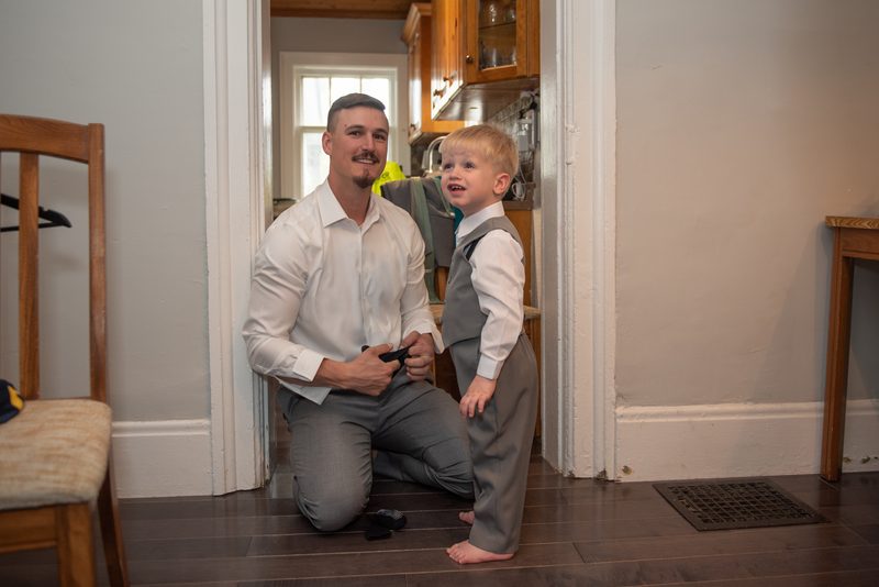 A groom kneels in a doorway adjusting his belt buckle while a young boy in matching attire stands beside him smiling.