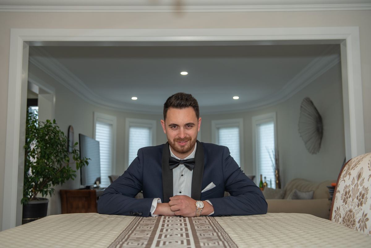 A man in a tuxedo smiles at the camera in an elegant dining room with soft lighting and natural light from windows.