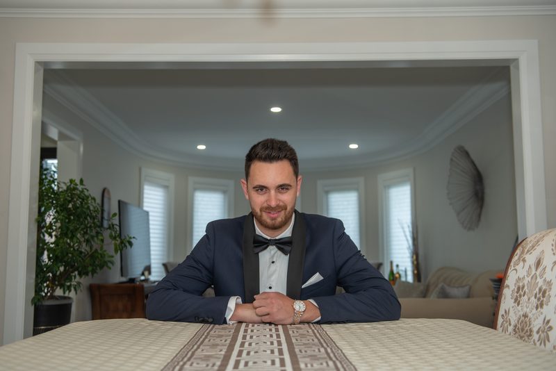 A man in a tuxedo smiles at the camera in an elegant dining room with soft lighting and natural light from windows.