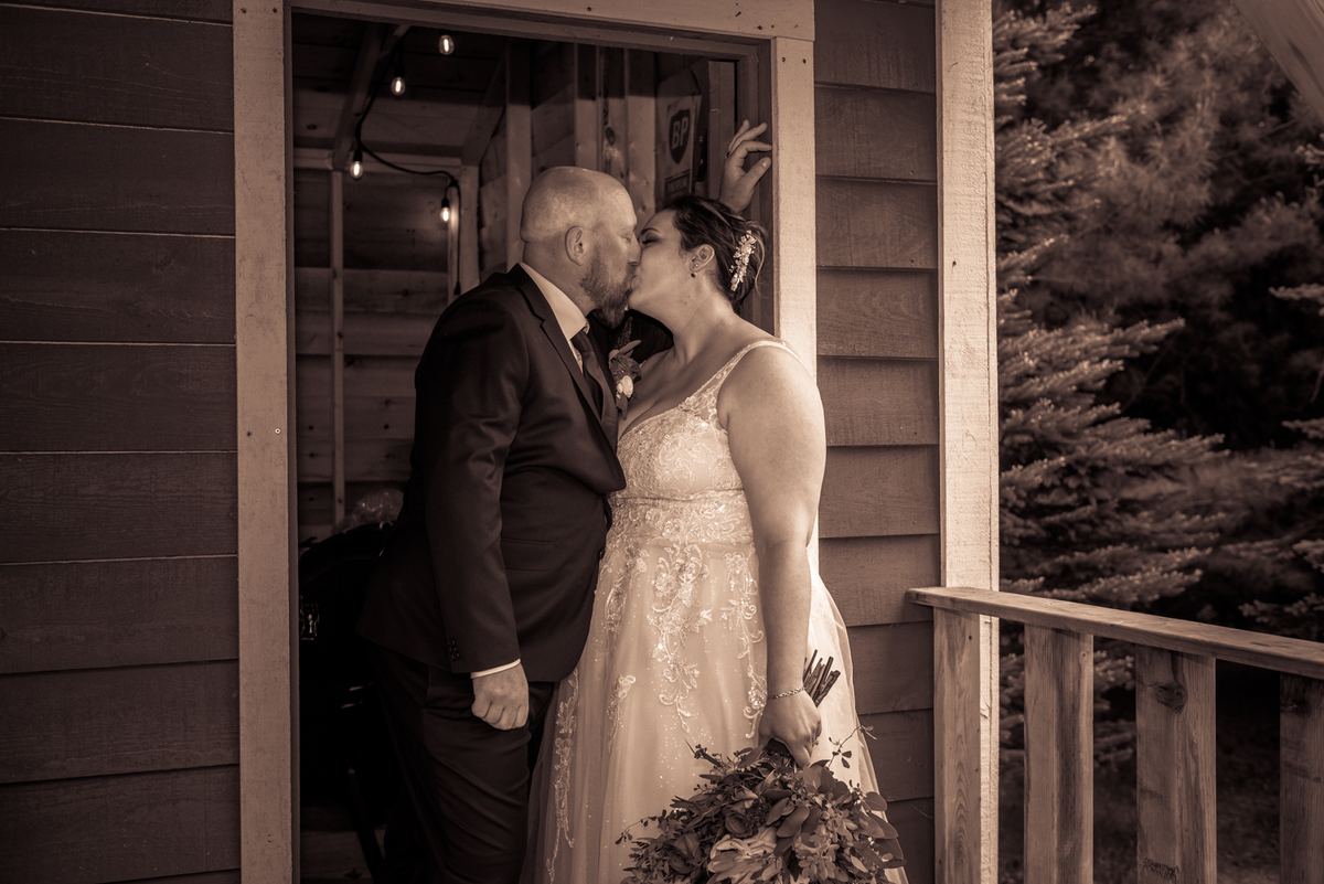 A newlywed couple kisses on a wooden porch at dusk, with warm string lights glowing inside the rustic cabin door frame.