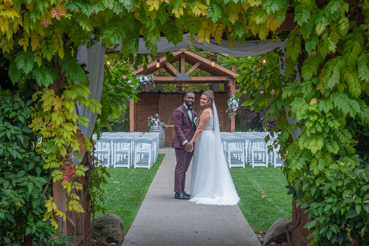 A smiling bride in white and groom in burgundy stand hand-in-hand at the end of a grassy aisle, framed by greenery.