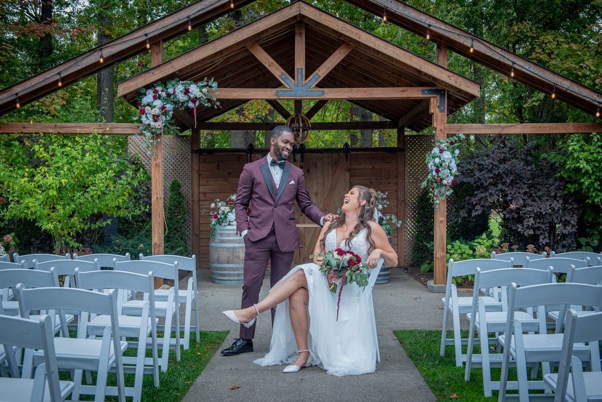A laughing bride and groom stand under a floral arbor adorned with lights, surrounded by white chairs.