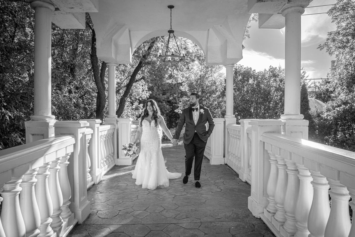 Bride and groom walk hand-in-hand along a sun-dappled veranda with white pillars and ornate railings.