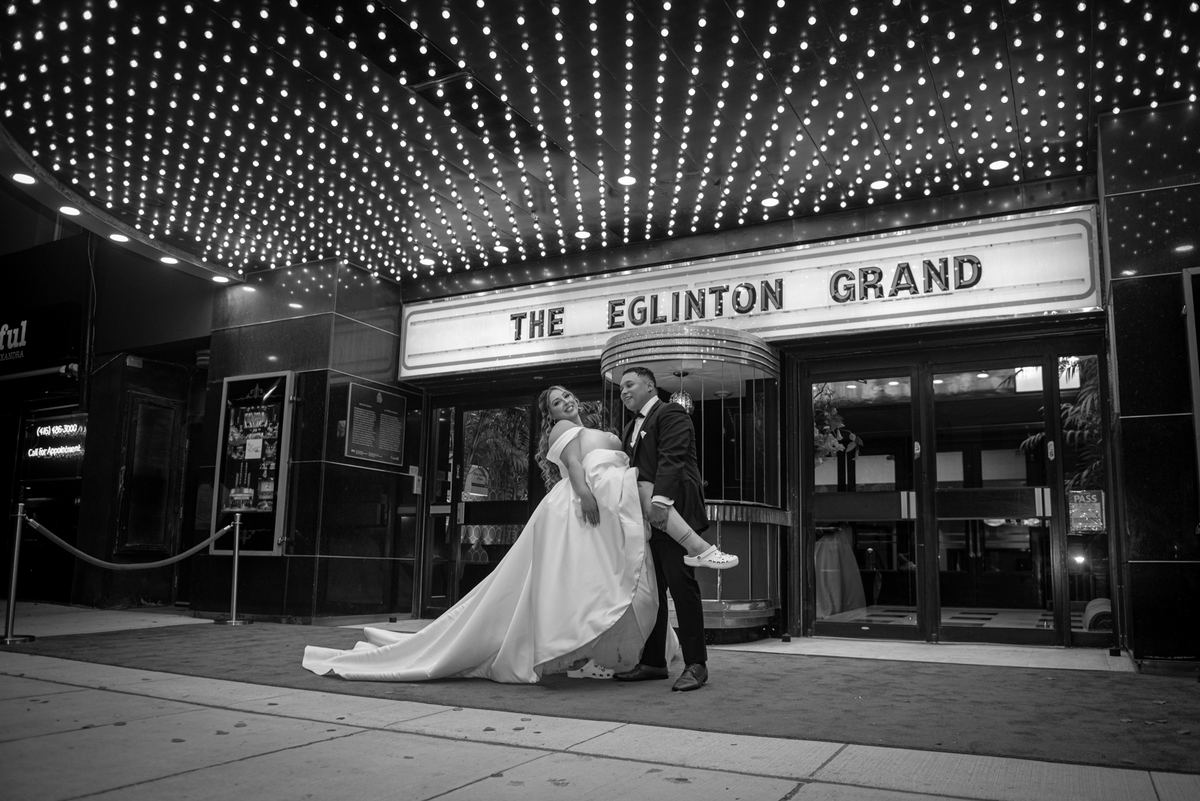 A bride and groom smile joyfully as the groom lifts her slightly, with string lights casting a glow over them.