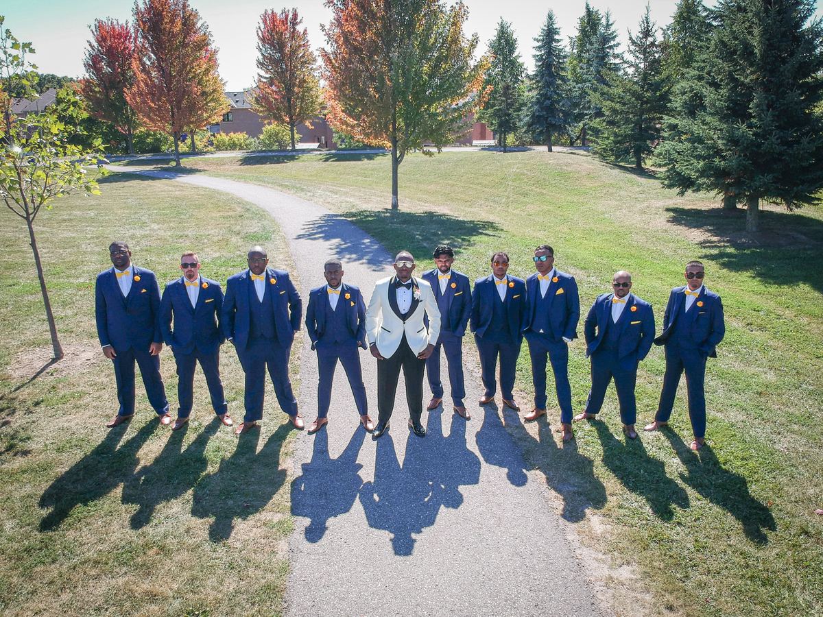 A groom and his groomsmen stand in navy suits with yellow bow ties on a park path, the sun casting long shadows.