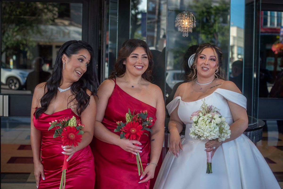 Bride in white and bridesmaids in red dresses smile with bouquets near glass doors reflecting street lights.