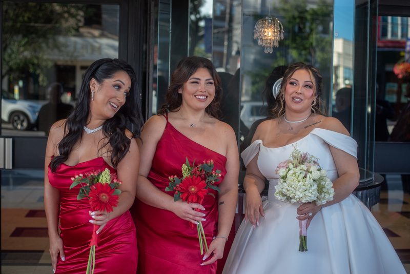 Bride in white and bridesmaids in red dresses smile with bouquets near glass doors reflecting street lights.