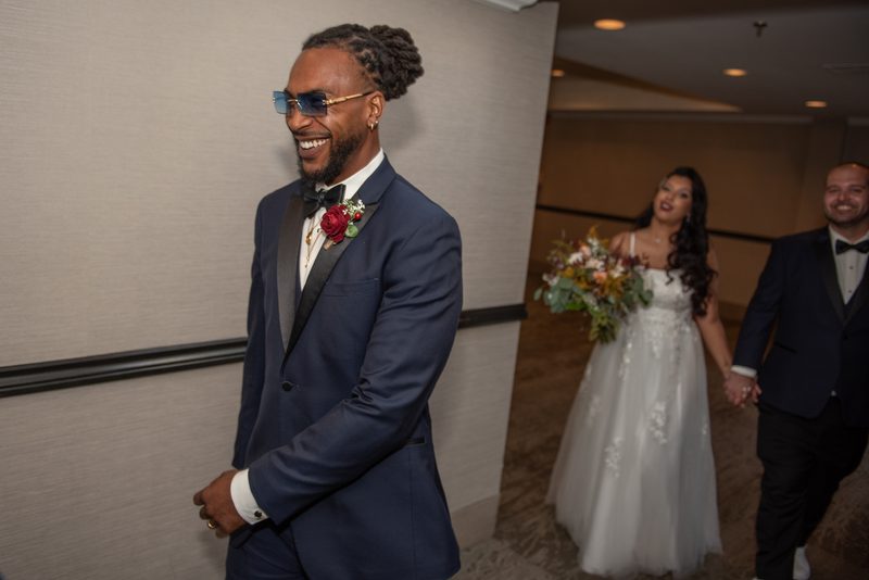 A groom in a navy tuxedo laughs as he walks with his bride and a groomsman, hand-in-hand, toward the ceremony.