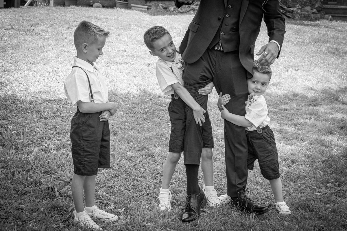 A man in formal wear surrounded by three young boys dressed formally, playfully interacting outdoors under dappled sunlight.