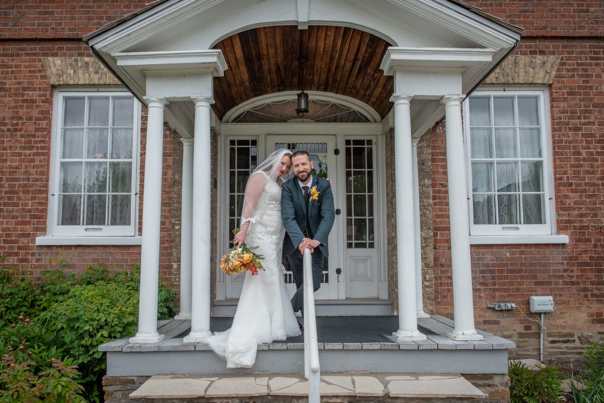 Jenna-Marie and Michael stand together on the porch steps of a brick house, smiling and leaning in affectionately.