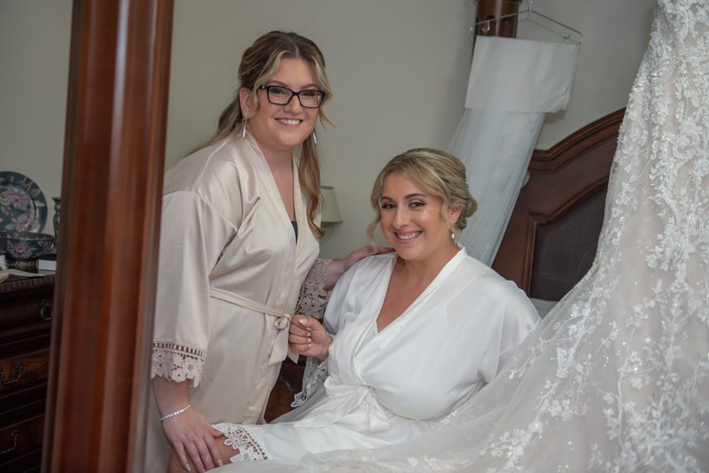 A bride in a white satin robe smiles as her friend adjusts the hem of her wedding gown hanging nearby, lace and veil visible.