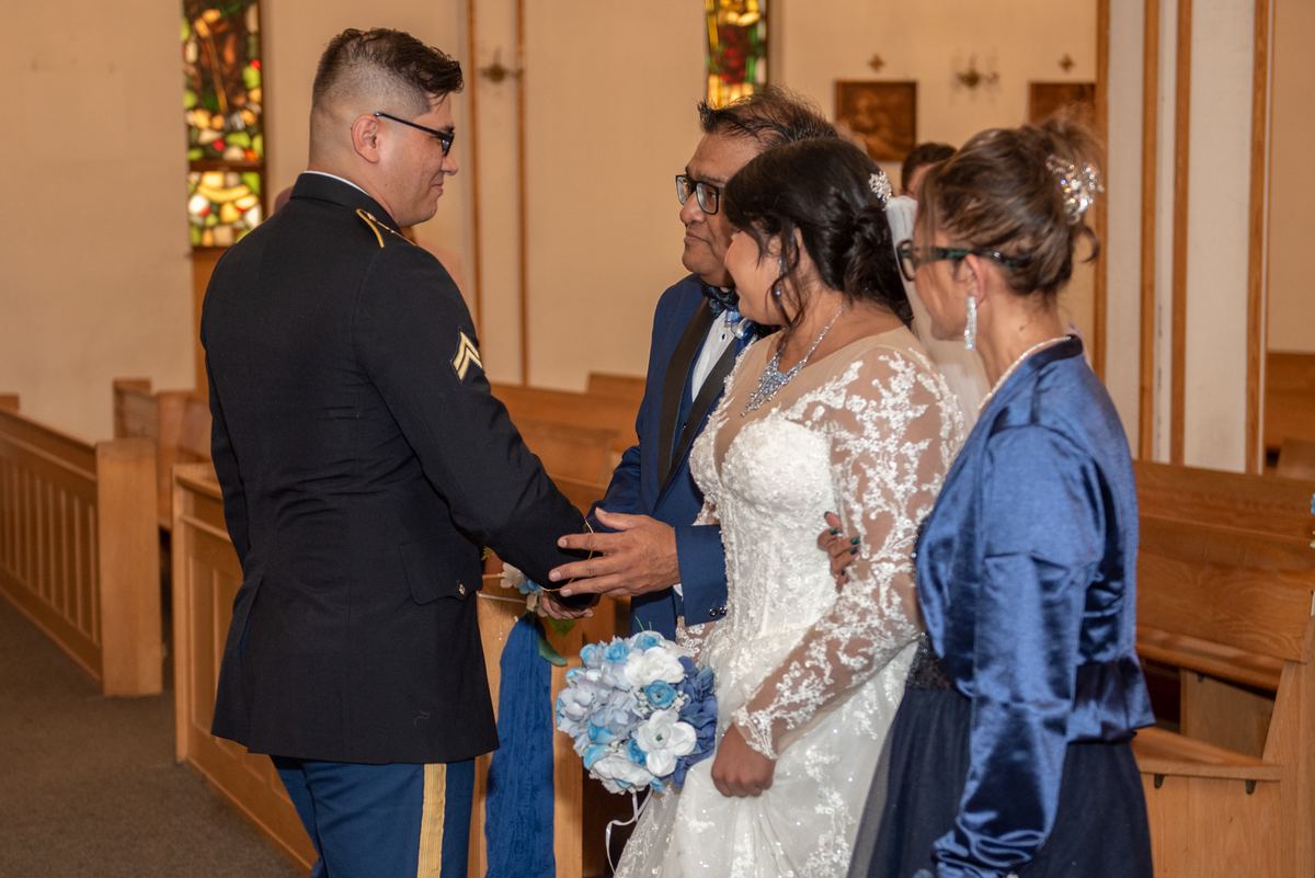 A bride in white and groom in uniform exchange vows, with stained glass windows casting light on wooden pews.